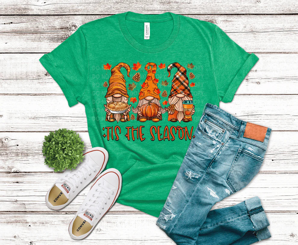 Green t-shirt with autumn-themed gnomes and 'Tis the Season text, paired with jeans and shoes on a wooden surface.