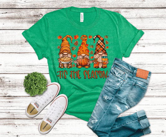 Green t-shirt with autumn-themed gnomes and 'Tis the Season text, paired with jeans and shoes on a wooden surface.